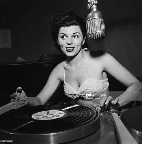 An unspecified female radio disc jockey, wearing a bikini top, smiling as she speaks into a microphone in an unspecified radio studio, location unspecified, location unspecified, United States, circa 1955